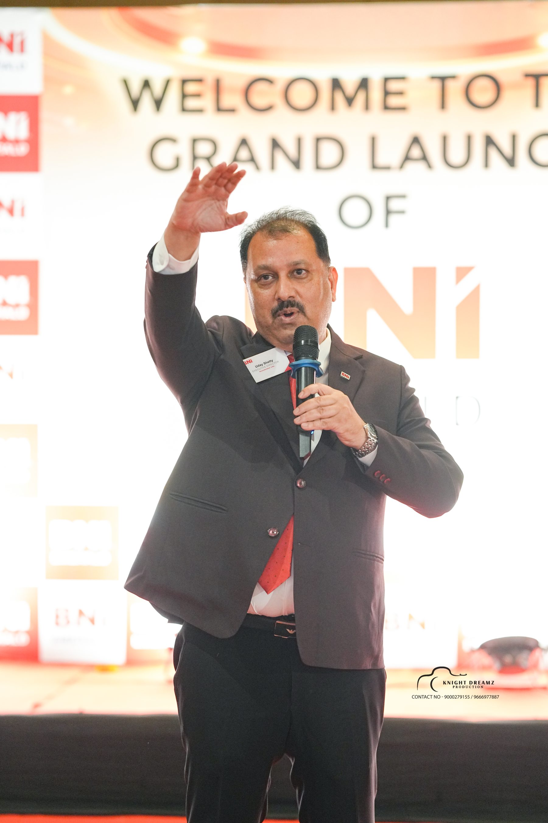 Uday Shetty gesturing on stage under Grand Launch backdrop